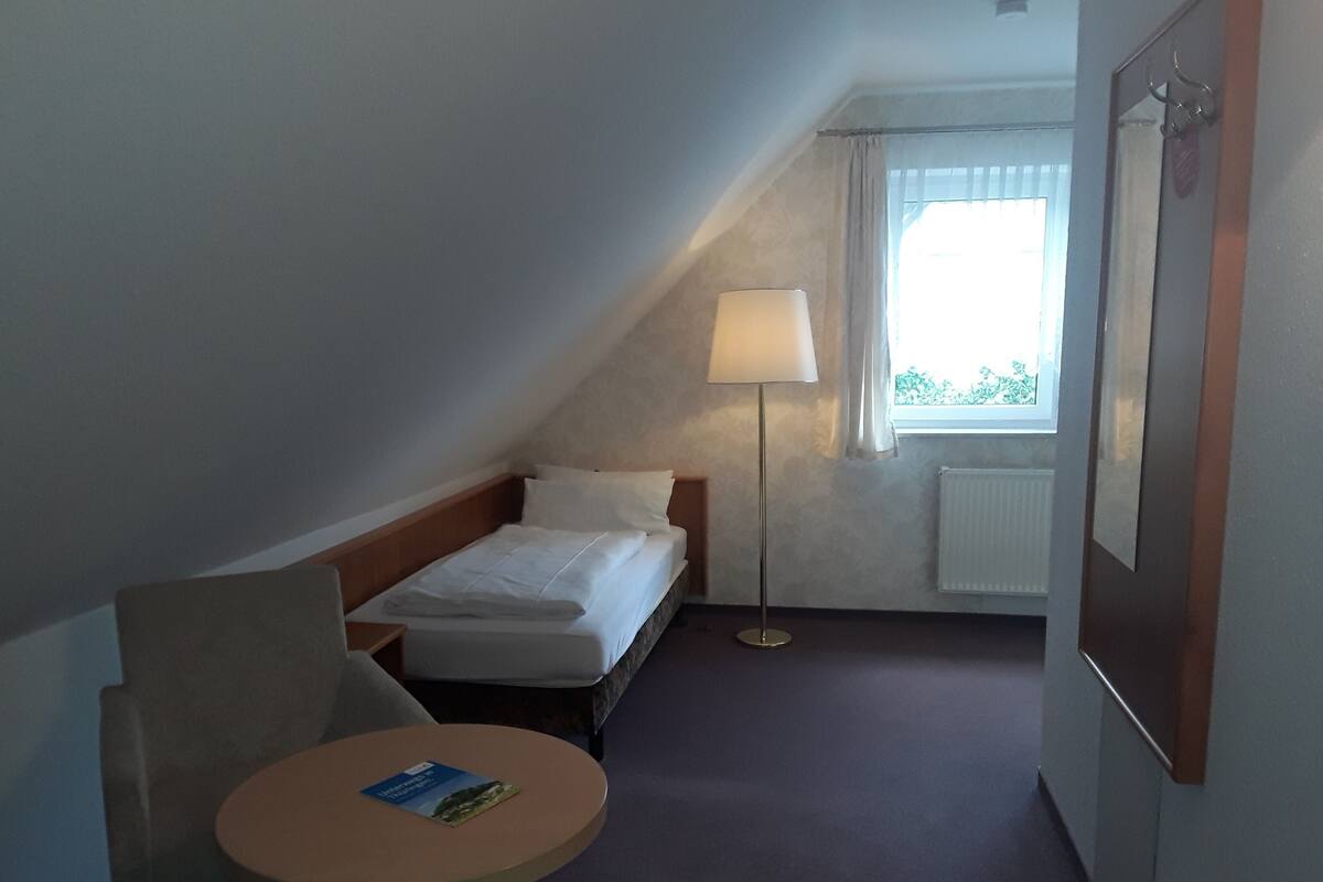 Una habitación pequeña, con poca luz, que tiene una cama individual, una mesita de noche con un libro, una lámpara y una ventana con cortinas.