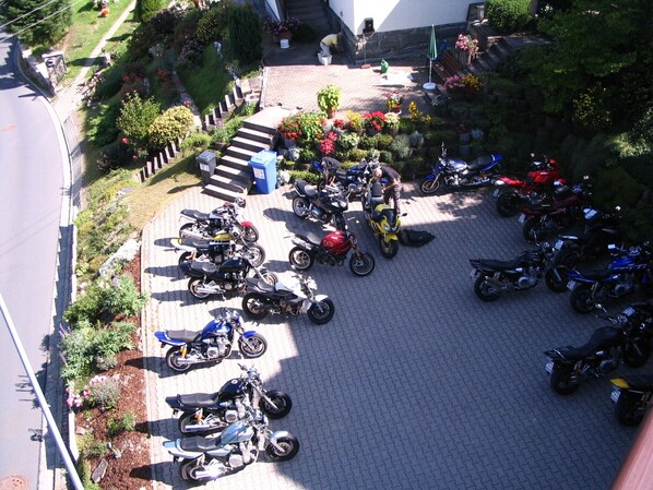 Un aparcamiento con varias motocicletas y ciclomotores, rodeado de vegetación y escalones que conducen a un edificio.