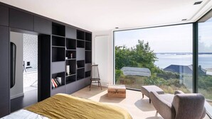 A modern bedroom with a large bed, a built-in bookshelf, a small stool, and a view of the sea through large windows. - To complete (Saint-Cast-le-Guildo)