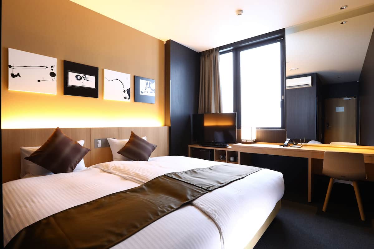 Ein Hotelzimmer mit einem großen Bett, einem Schreibtisch und einem Fernseher. An der Wand über dem Bett befinden sich Kunstwerke und ein Fenster mit Vorhängen.