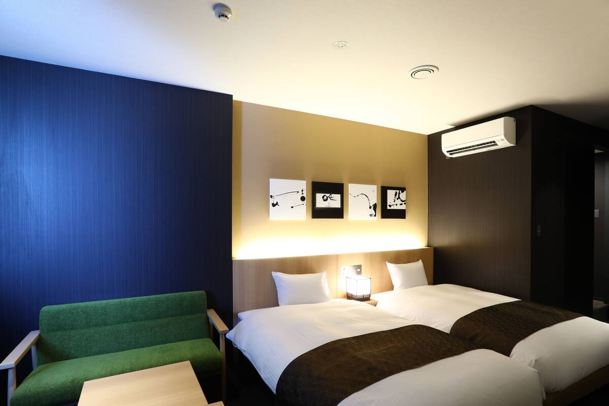 Ein Hotelzimmer mit einem Bett, zwei Nachttischen, einem grünen Sessel und einer Wand-Klimaanlage.