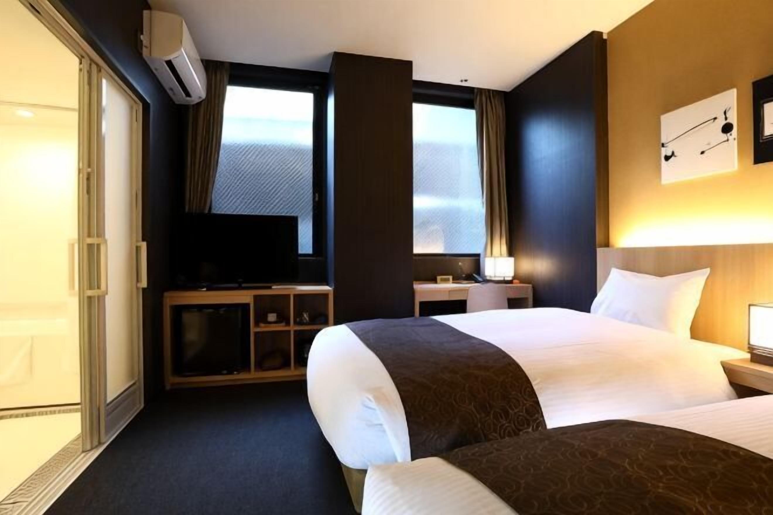Quarto de hotel com duas camas, televisão, escrivaninha e uma janela grande.