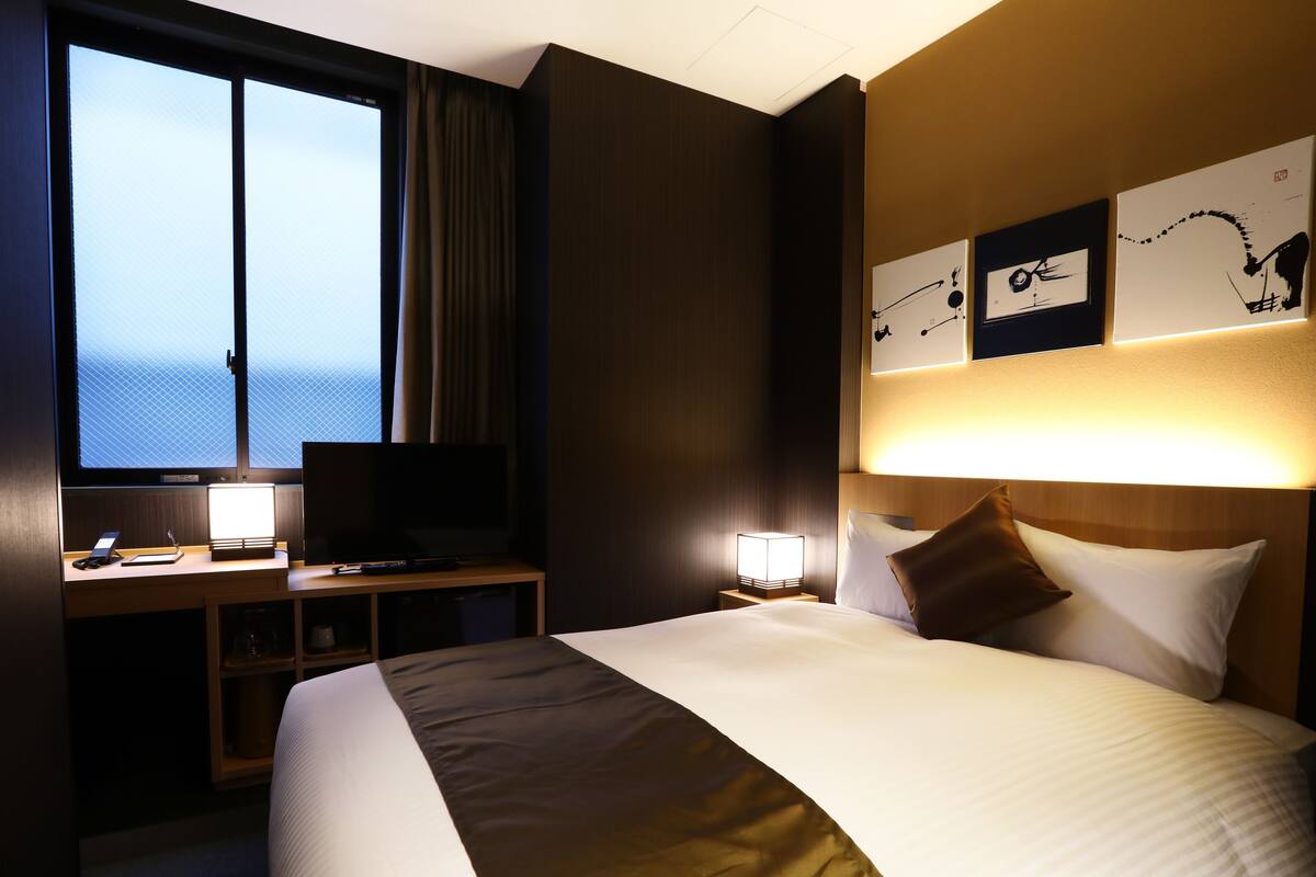 Ein Hotelzimmer mit einem großen Bett, einem Schreibtisch mit Computer und Fernseher, einem Fenster mit Vorhängen und Kunstwerken an der Wand.