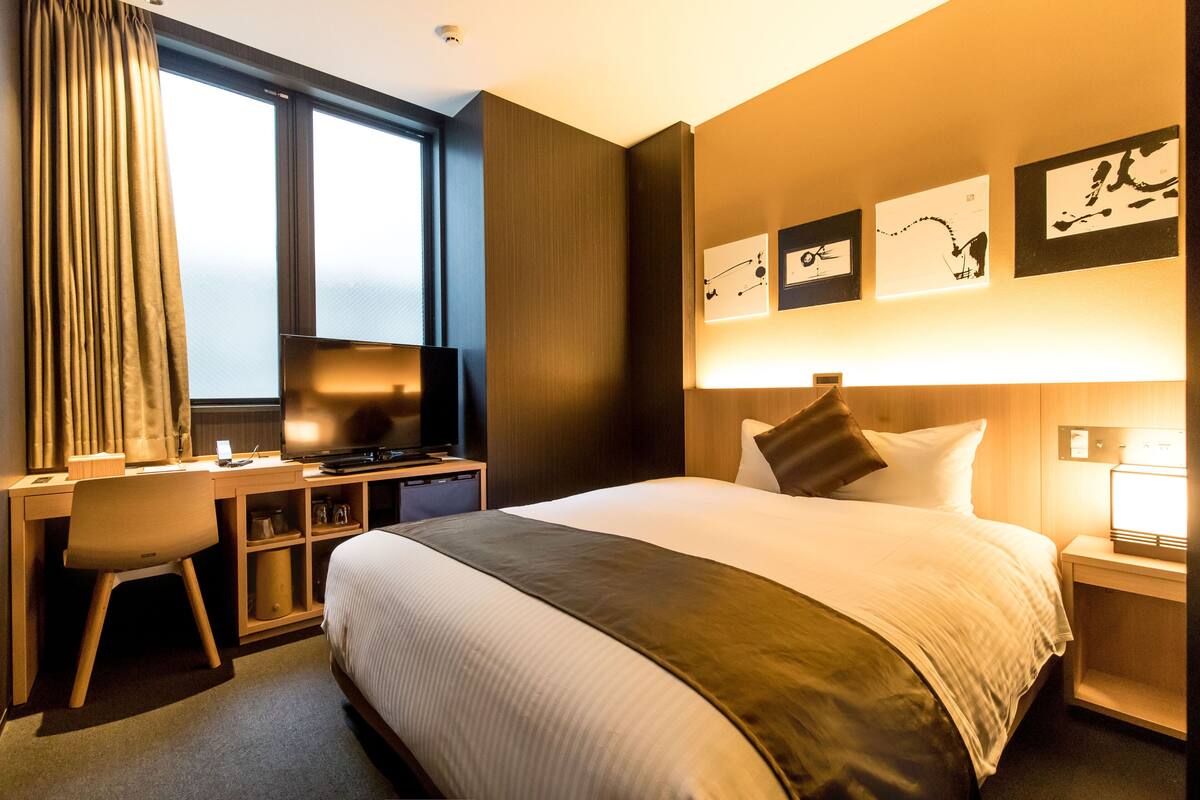 Ein Hotelzimmer mit Bett, Schreibtisch, Stuhl, Fernseher, Wandbild und einem Fenster mit Vorhängen.