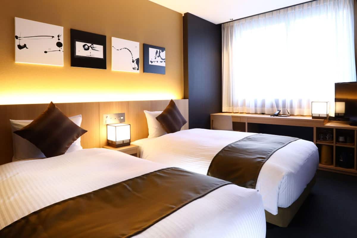 Ein Hotelzimmer mit zwei Betten, einem Schreibtisch mit Fernseher, einer Lampe und einem Kunstwerk an der Wand.