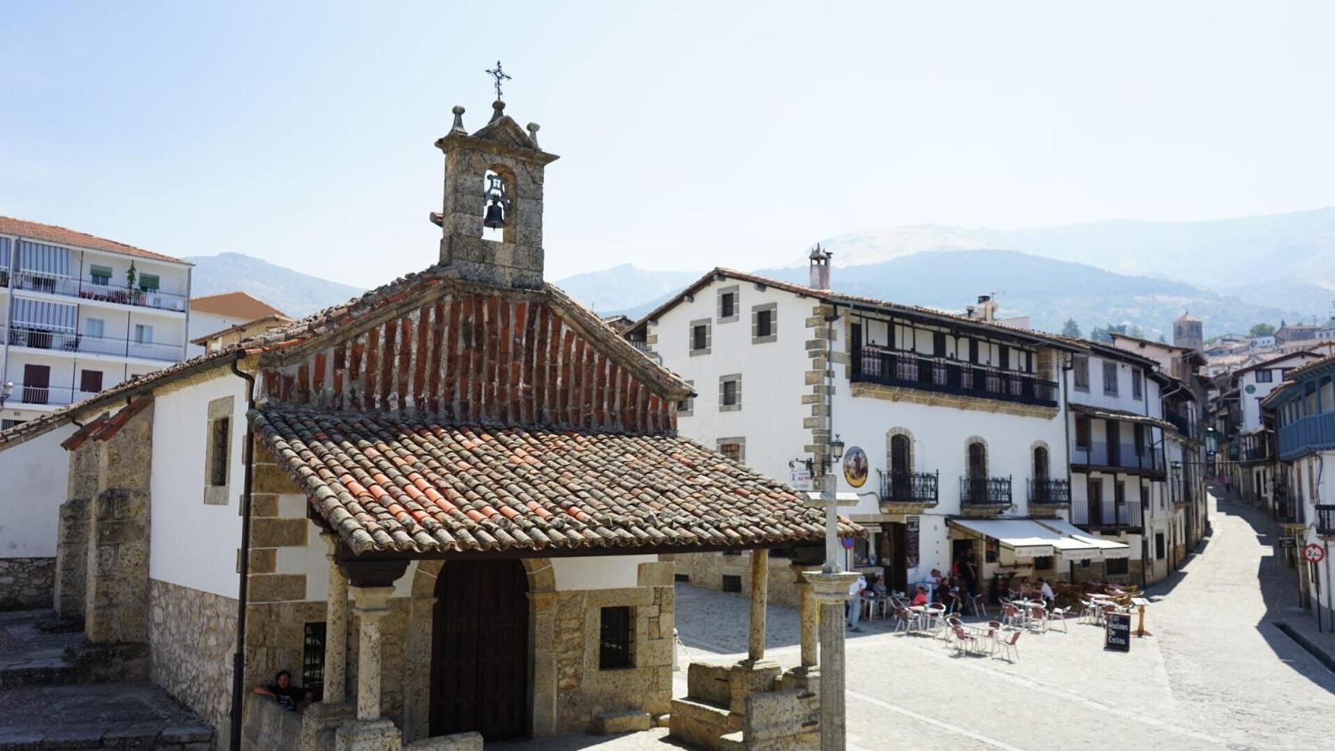 Una iglesia histórica con un campanario, un patio con mesas al aire libre y edificios tradicionales en el fondo.