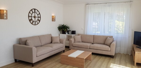 Ett vardagsrum med två beige soffor, ett soffbord i trä, ett TV-bord och ett stort fönster med tunna, genomskinliga gardiner.
