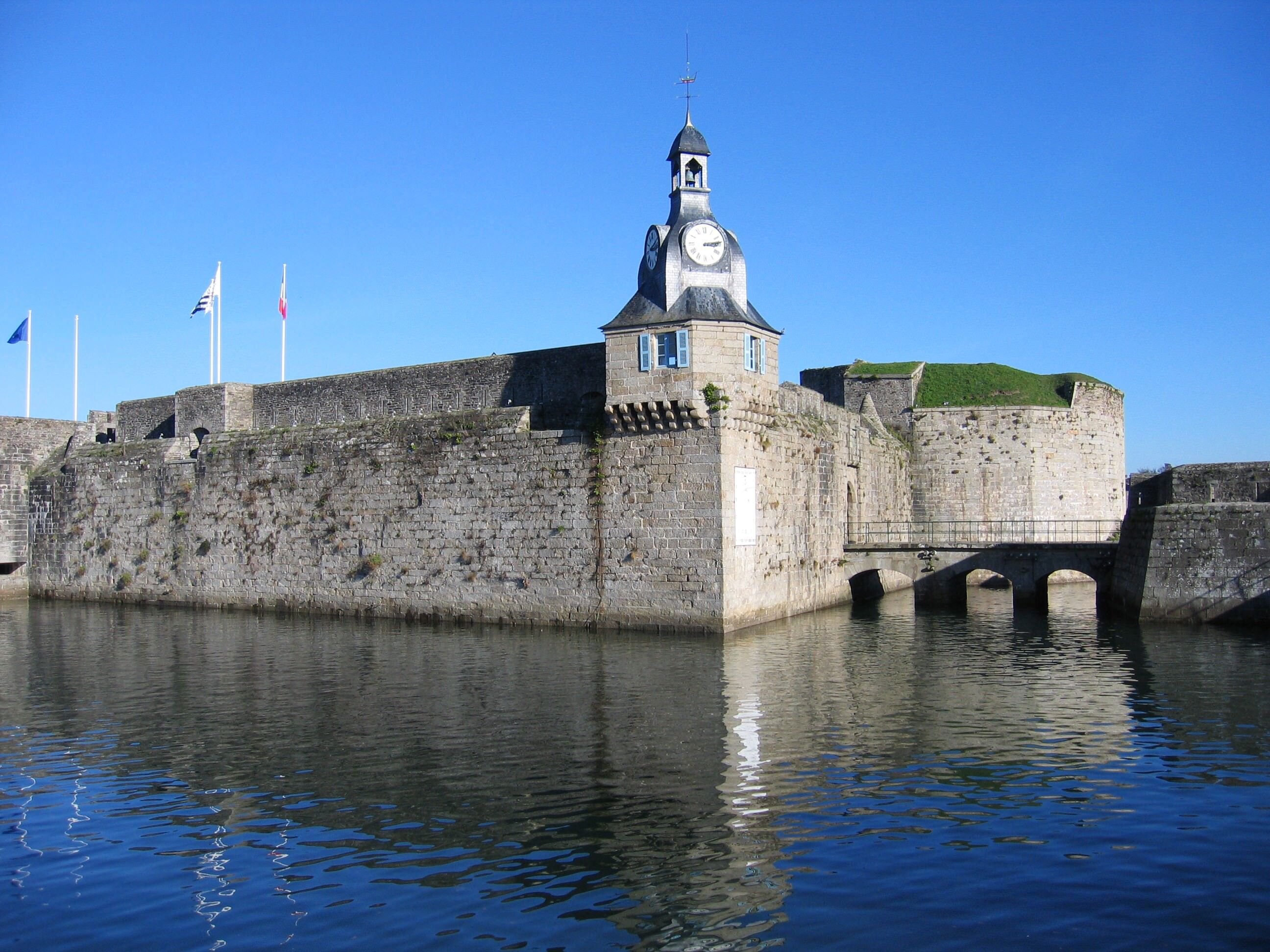 A historic stone fortification with a clock tower, a bridge, and flags.