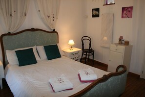 A neatly made bed with a wooden headboard, two pillows, and two folded towels. There is a nightstand with a lamp, a chair, and a small cabinet with a mirror.