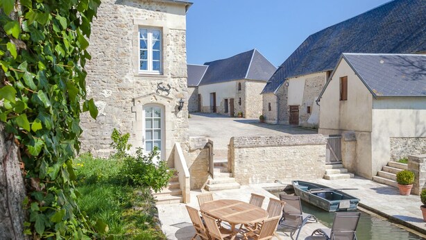 A stone building with a window and a door, a paved patio with a table and chairs, and a small body of water. - Luxury Cottage, Outdoor Nordic Hot Tub and Sauna, D-Day Beaches 15 km (Isigny-Sur-Mer)