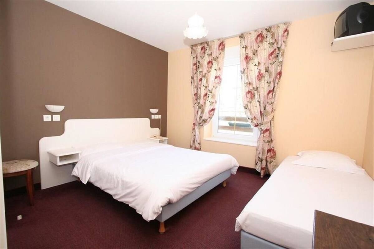 A bedroom with a single bed, a bedside table, a chair, a window with floral curtains, and a wall-mounted lamp.
