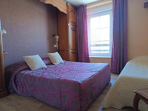 Ein Zimmer mit einem Bett, einem Holzschrank, einem Nachttisch, einem Fenster mit Vorhängen und einer Tür.