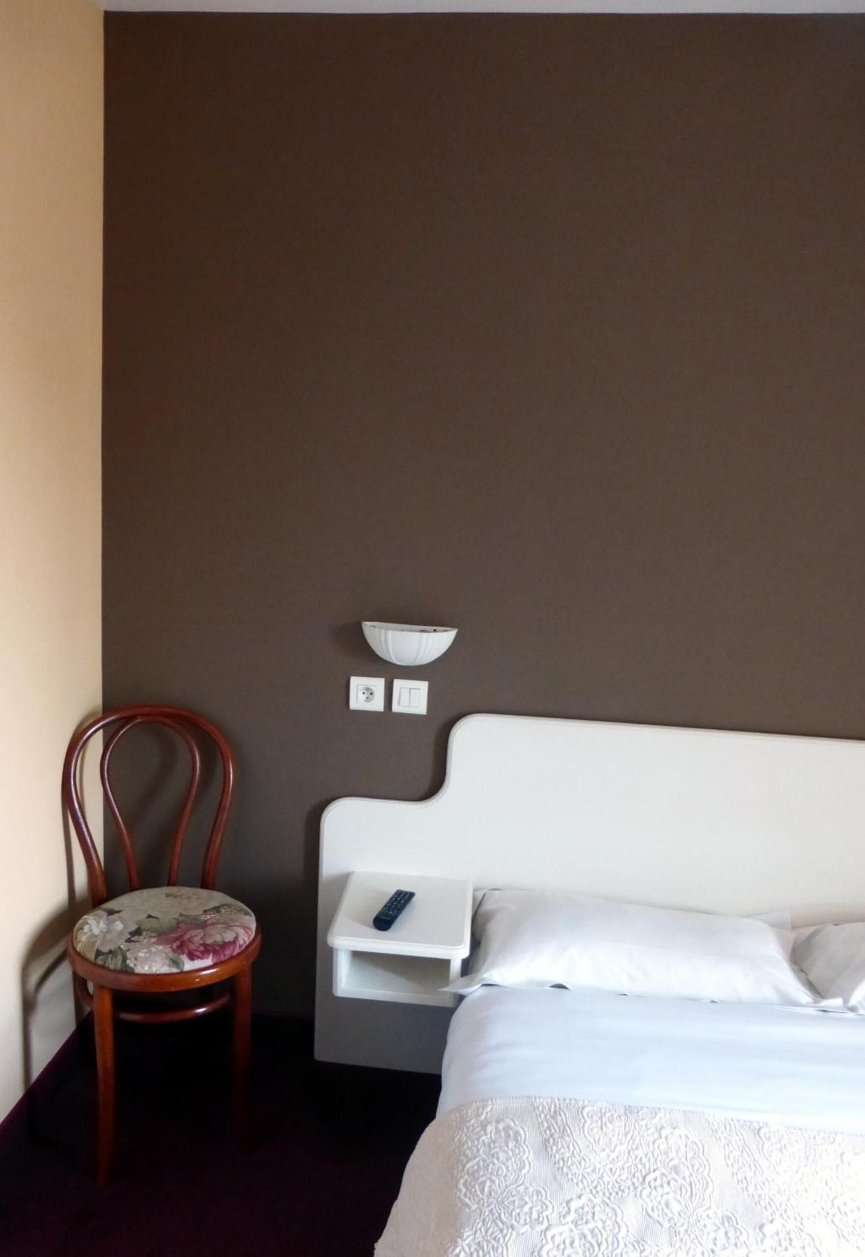A hotel room with a bed, a chair, a bedside table, and a wall-mounted light fixture.