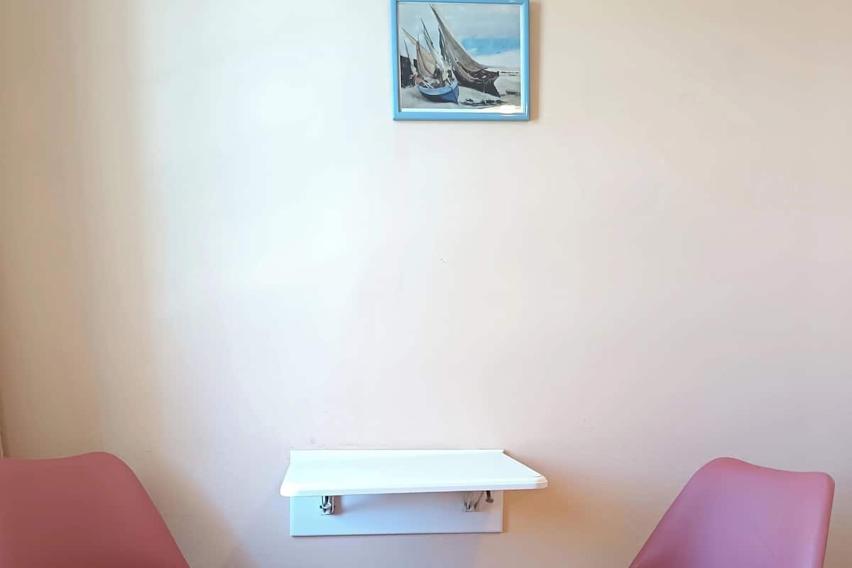 Two pink chairs, a white shelf, and a painting of boats on a wall.