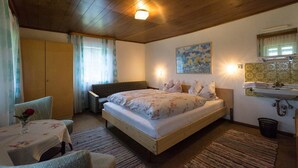 A bedroom with a wooden ceiling, a bed with floral bedding, a bedside table with a vase, a chair, a sofa, a painting on the wall, and a bathroom with a sink and mirror. - Haus am Waldesrand (Regen)