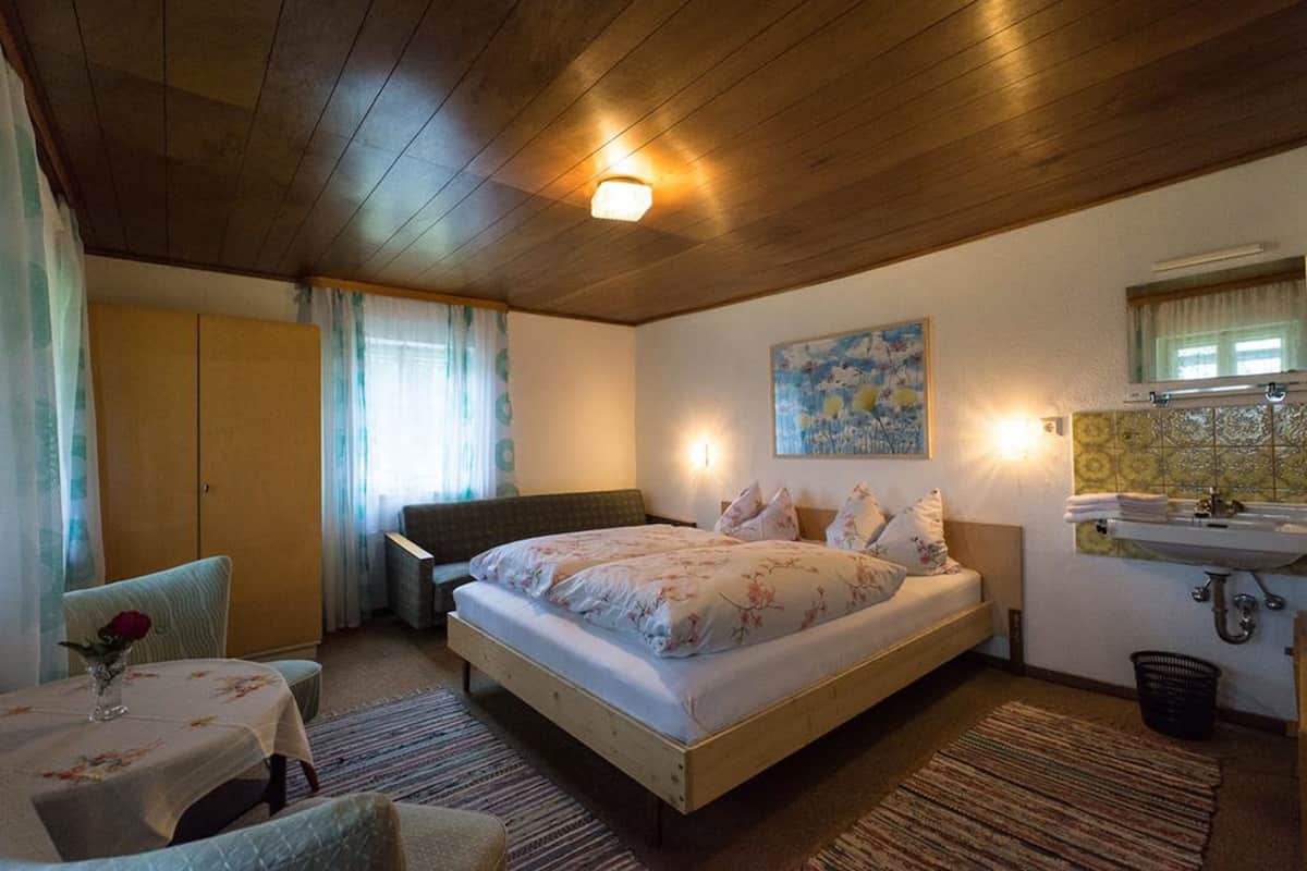 Un dormitorio con techo de madera, una cama con ropa de cama floral, una mesita de noche con un jarrón, una silla, un sofá, un cuadro en la pared y un baño con lavamanos y espejo.
