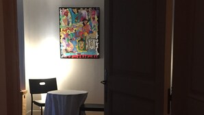 Ein Zimmer mit rotem Teppich, einem Tisch mit weißer Tischdecke, einem schwarzen Stuhl und einem farbenfrohen Kunstwerk an der Wand.