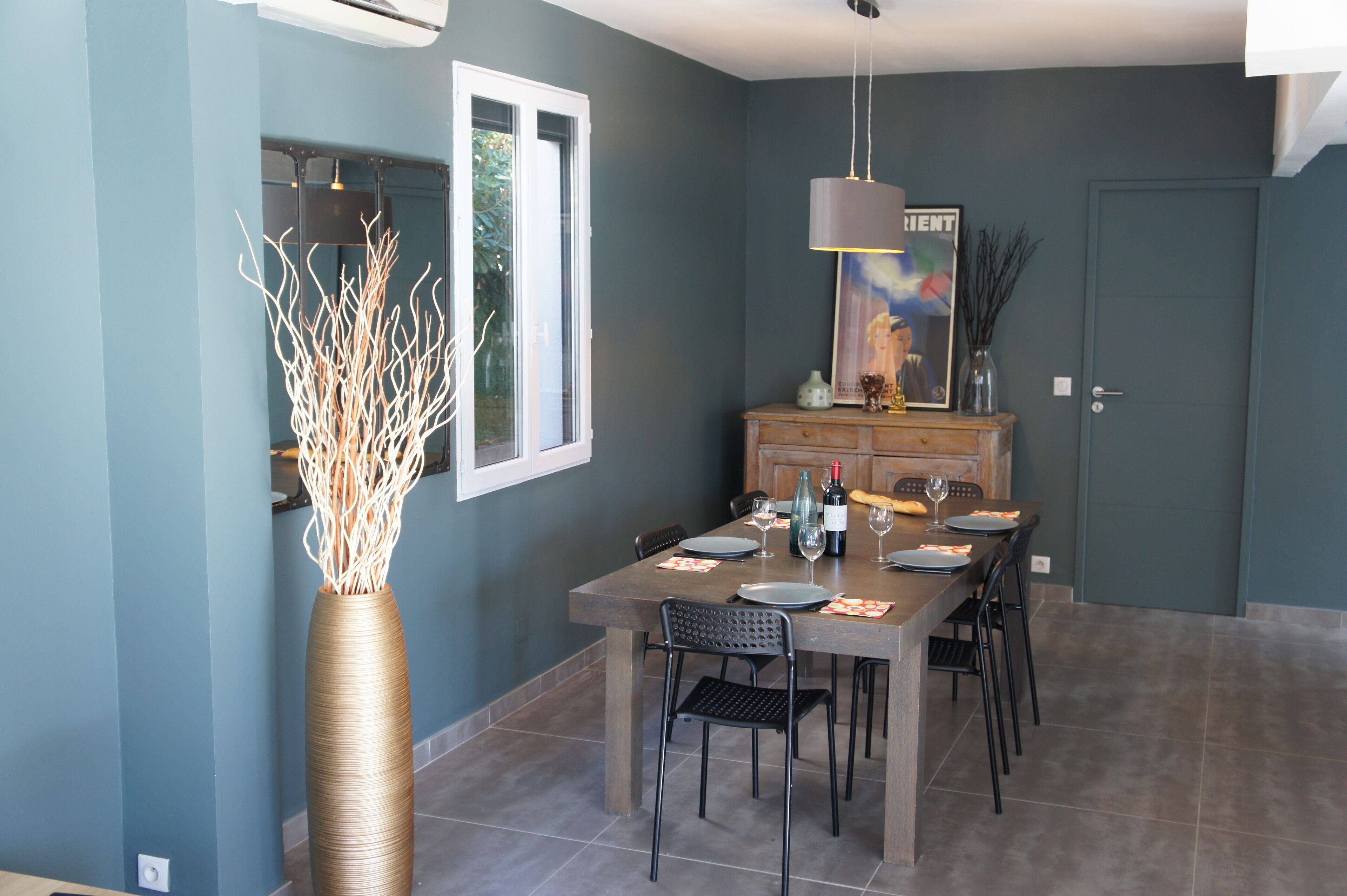 A dining area with a table set for six, a tall vase with branches, and a painting on the wall.