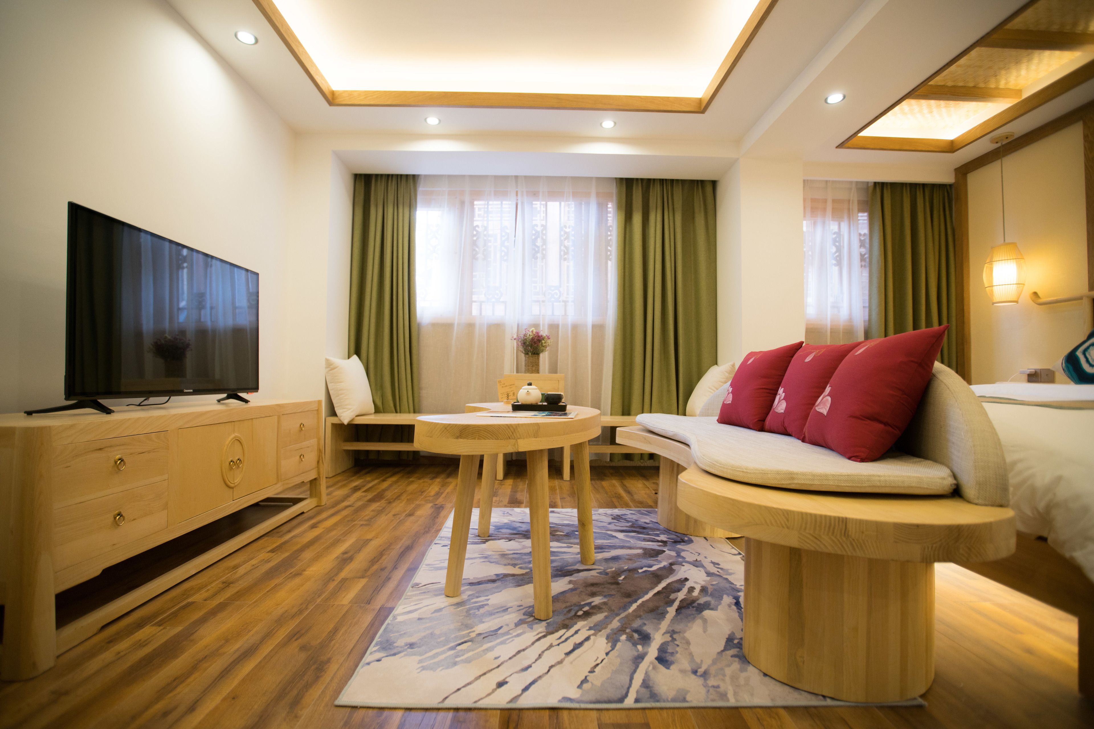A modern hotel room with a wooden floor, a flat-screen TV, a round wooden table, a sofa with red cushions, and a bed with a white duvet.