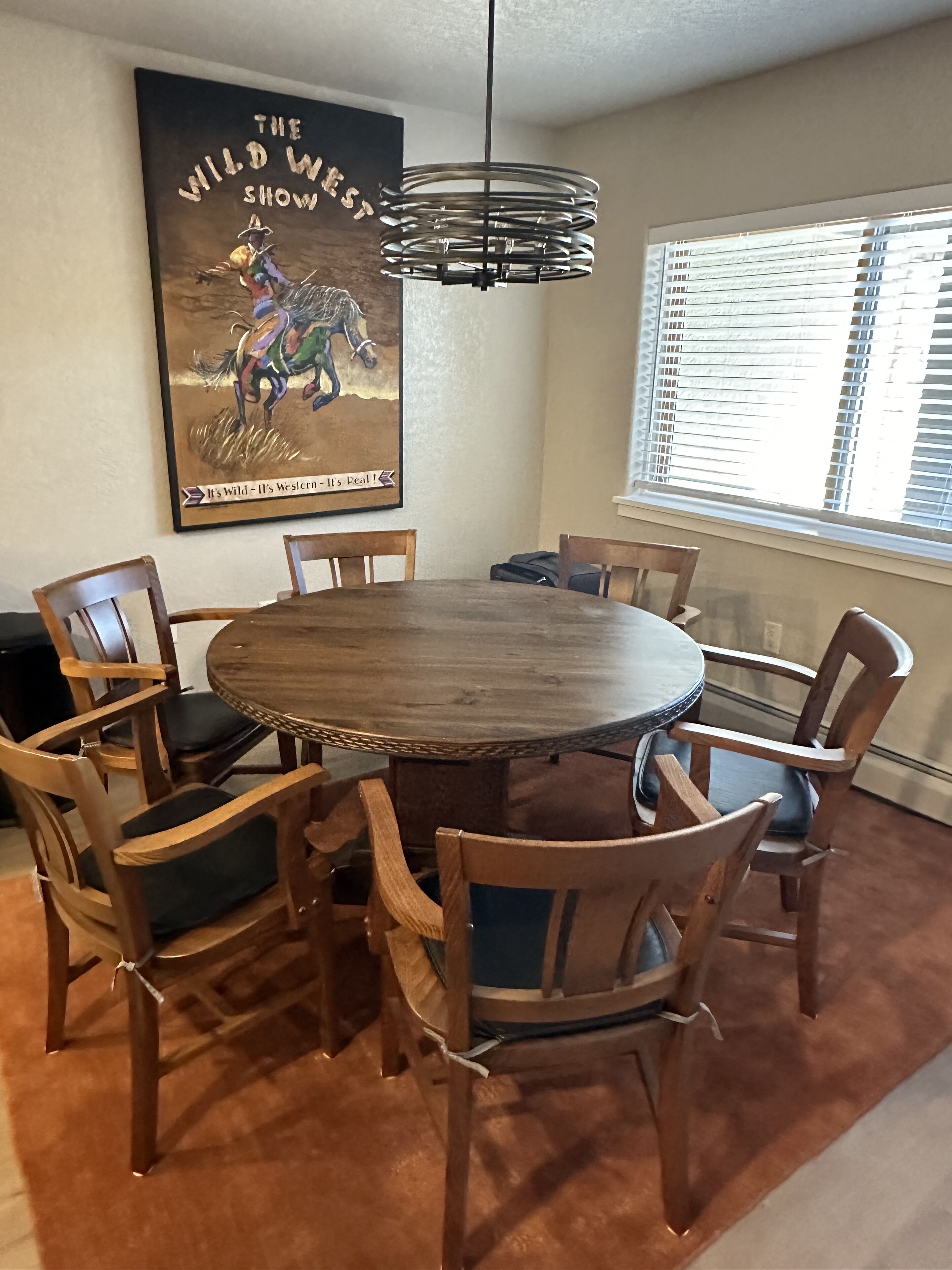 A dining area with a round wooden table surrounded by six wooden chairs with cushioned seats. A large poster of a cowboy on a horse is mounted on the wall above the table.