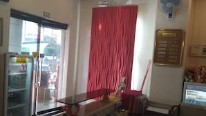 Zona d'entrada d'hotel amb un taulell de recepció, un ventilador, un panell informatiu fixat a la paret i una finestra amb vista a l'exterior.