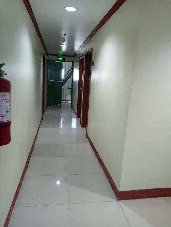 Corredor limpo e bem iluminado, com um extintor de incêndio à esquerda, uma escada ao centro e uma porta à direita.