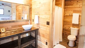 Baño de cabaña de madera con lavamanos de madera, espejo, inodoro y zona de ducha.