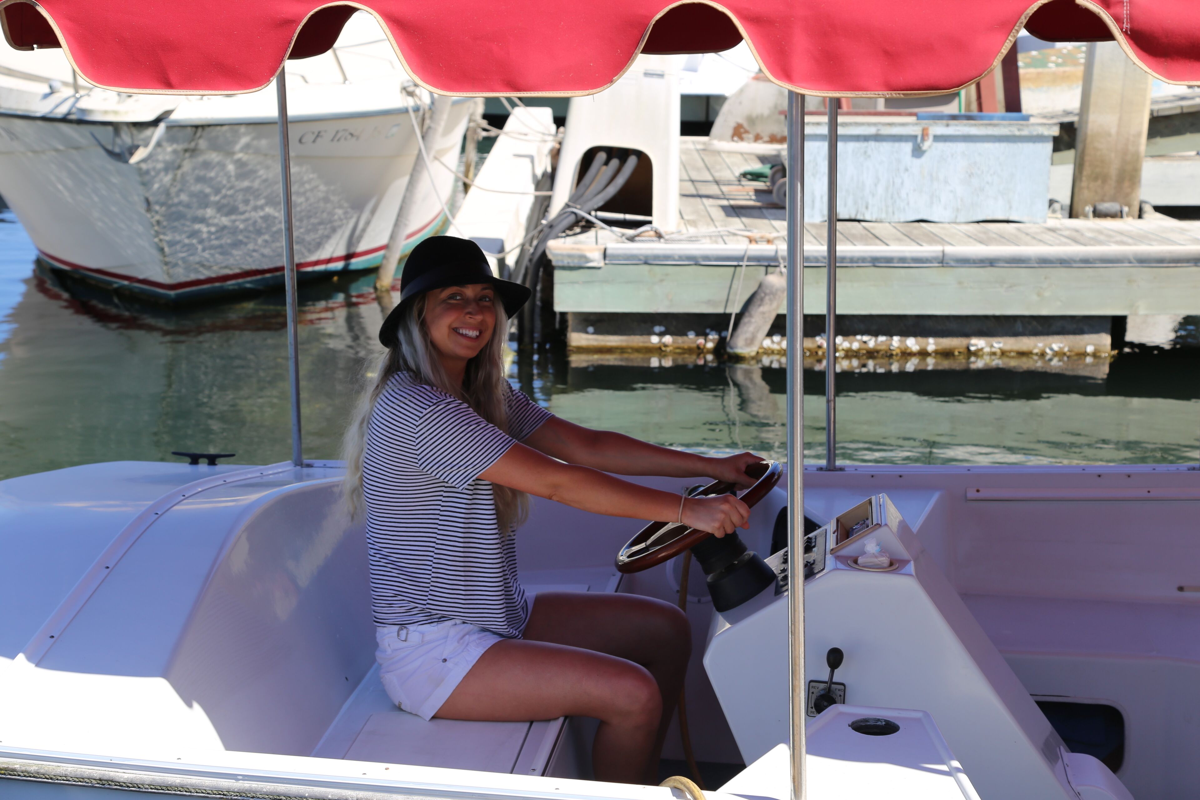 Une personne qui tient la barre d’un bateau, un auvent rouge la protégeant du soleil.