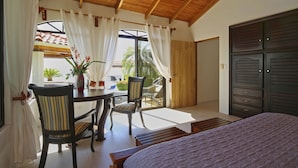 Ein Schlafzimmer mit Bett, Holzboden, einem Fenster mit Vorhängen, einem Tisch mit Stühlen und einer Zimmerpflanze.