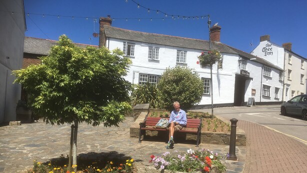 A man sitting on a bench in a flower bed with a white building in the background. - The Tree Inn (Bude)