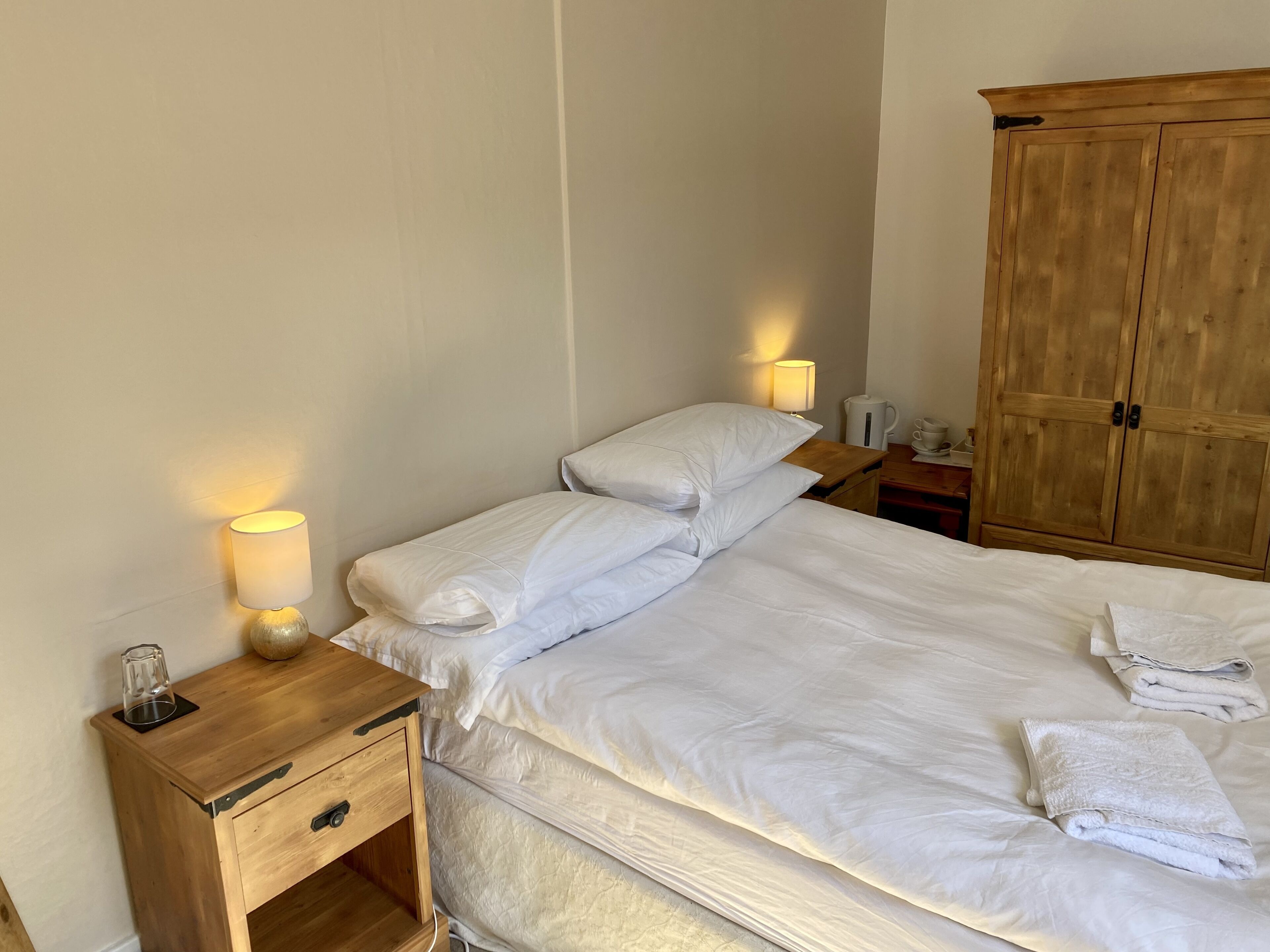 Een net opgemaakt bed met witte lakens, een bedlampje, een houten nachtkastje en een houten garderobe.