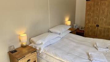 Een net opgemaakt bed met witte lakens, een bedlampje, een houten nachtkastje en een houten garderobe.