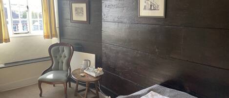 A hotel room with a dark wooden wall, a framed picture, a chair, a small round table with a kettle, and a bed with a folded towel.