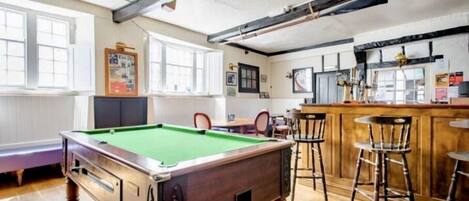 A billiards table in a room with wooden floors, a bar area with stools, and a window with blinds.