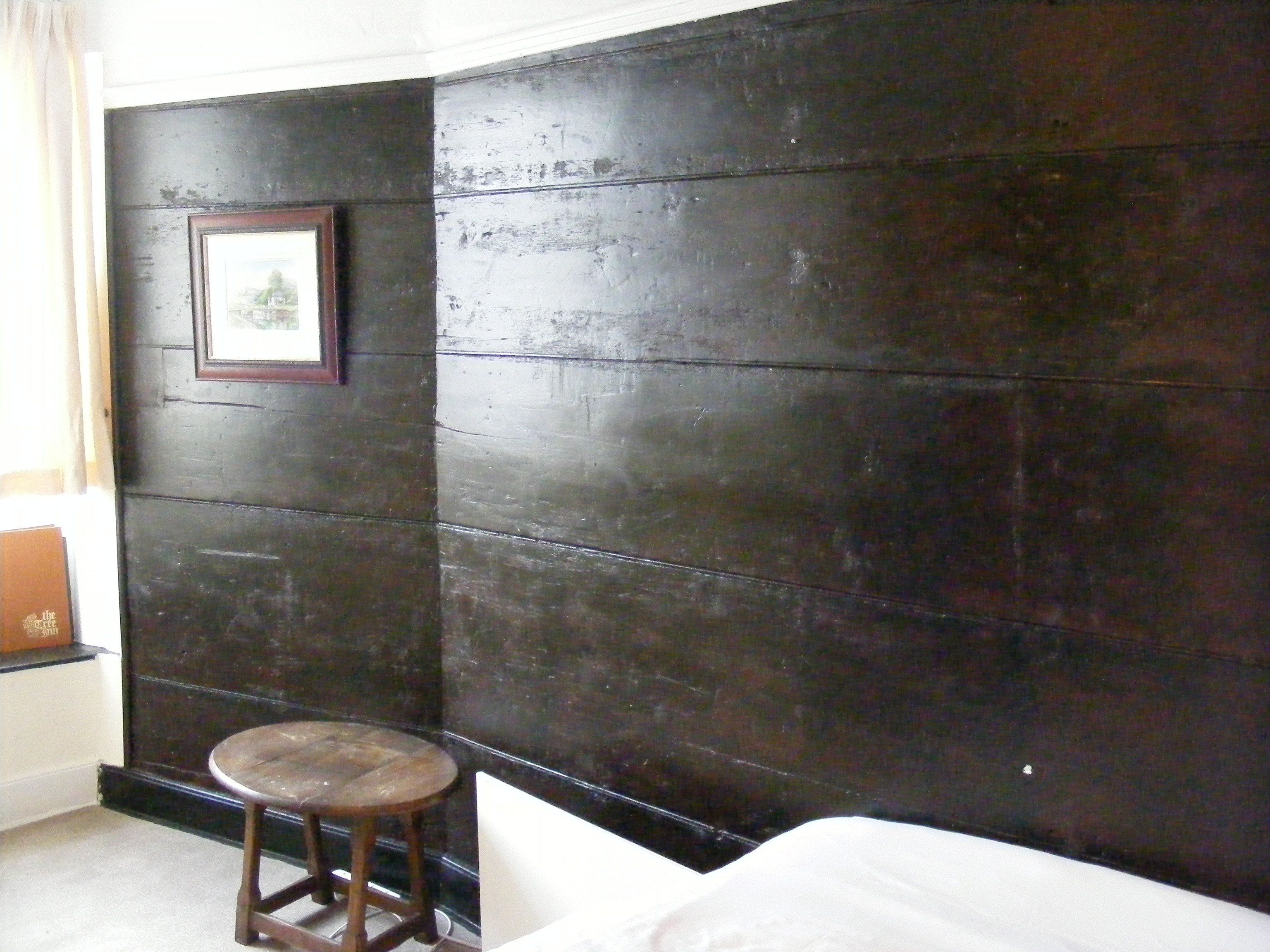 A room with a dark wooden wall, a framed picture, a small wooden stool, and a bed with white bedding.