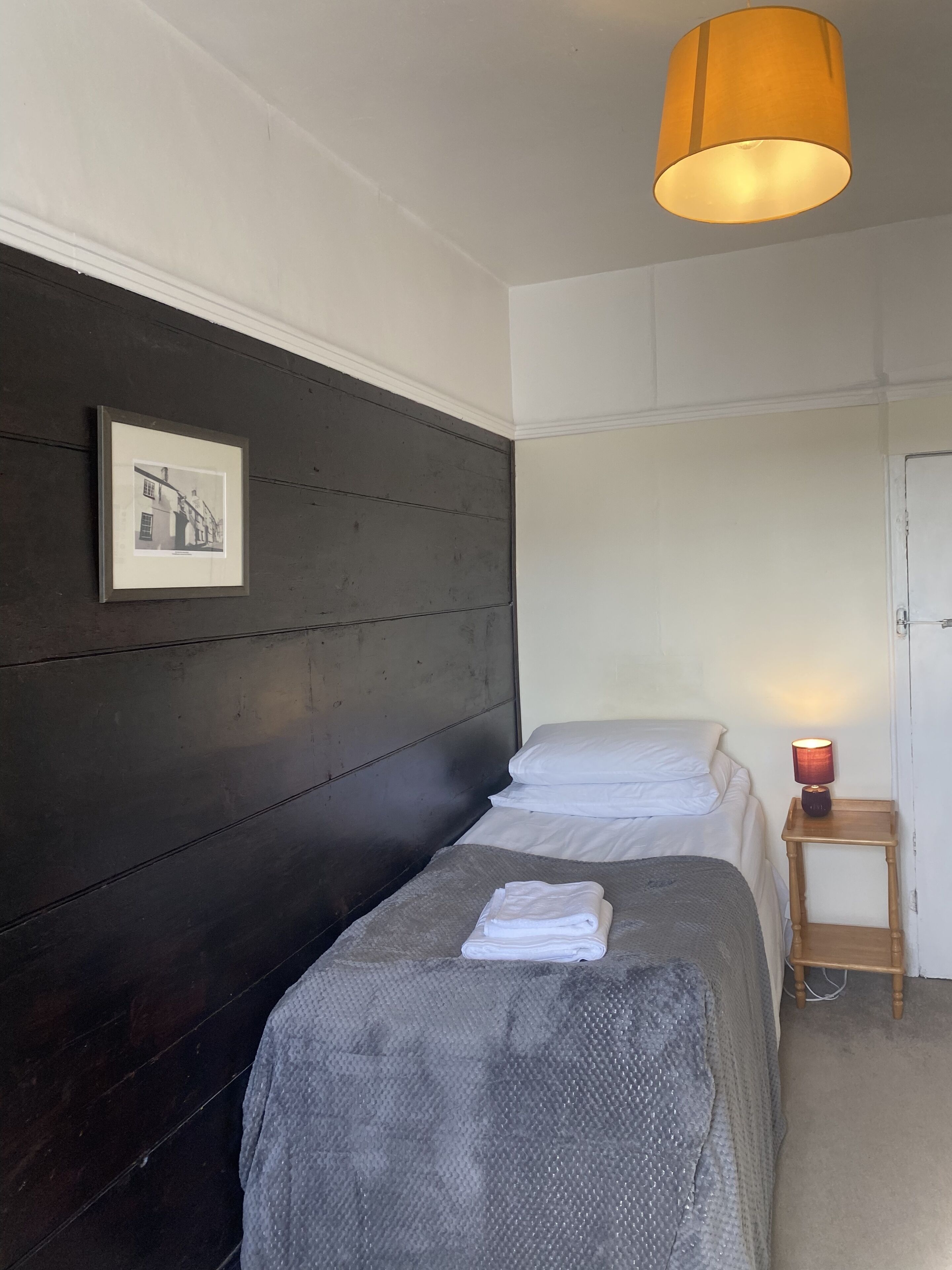 Une chambre avec deux lits, un tableau encadré au mur, une lampe sur une table de chevet et une porte sur le côté droit.