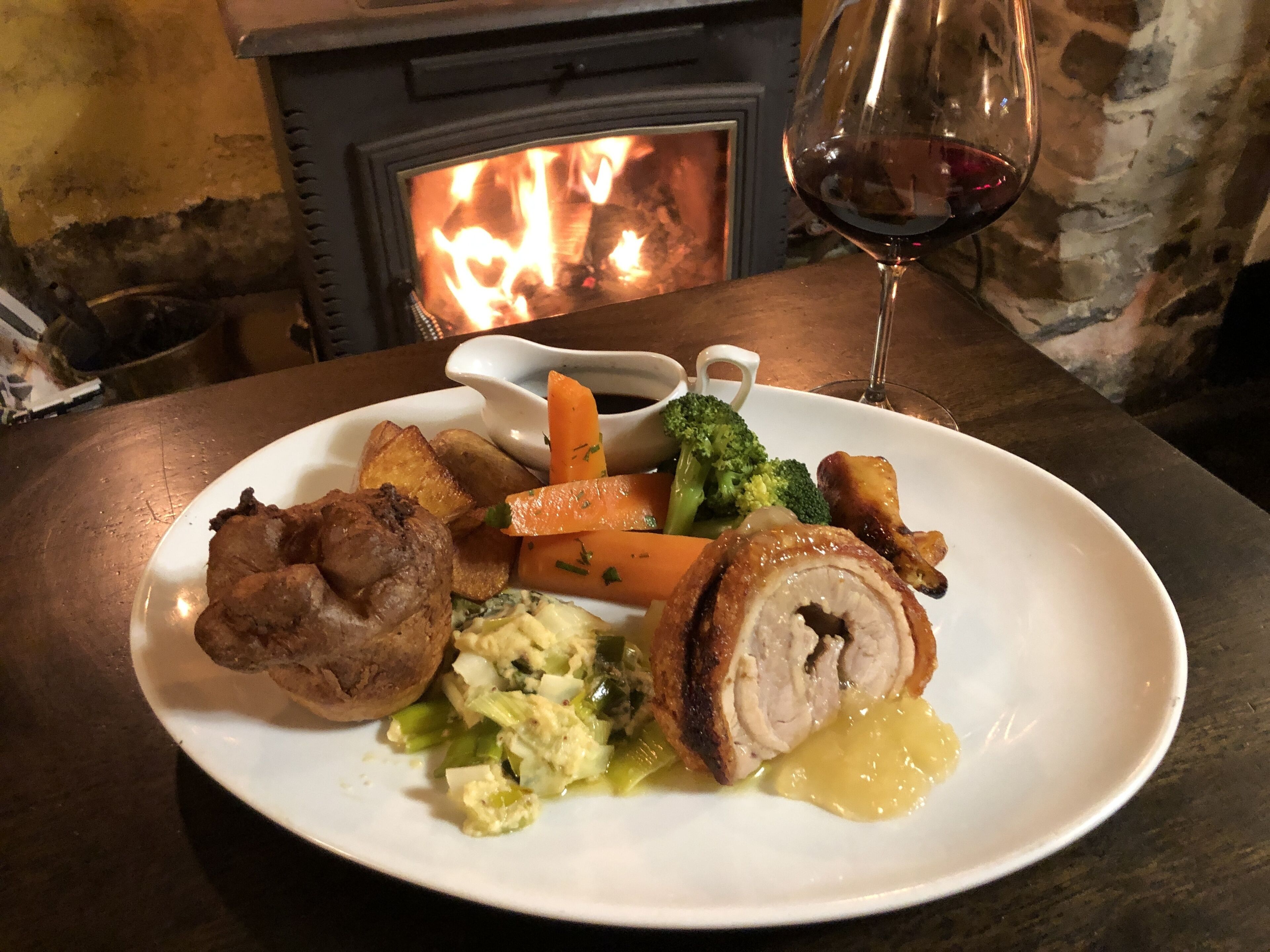 A plate of food with roasted meat, vegetables, and gravy, accompanied by a glass of red wine, set on a table with a fireplace in the background.