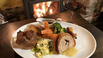 A plate of food with roasted meat, vegetables, and gravy, accompanied by a glass of red wine, set on a table with a fireplace in the background.