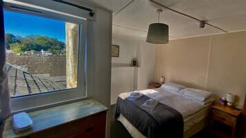 Un dormitorio con cama, cómoda de madera, mesita de noche, lámpara y ventana con vista a árboles y edificios.