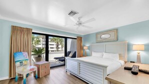 A hotel room with a bed, a chair, a desk, a ceiling fan, and a view of palm trees through the window. - Aegean 215 – Updated Condo with Private Balcony & Beach Access (Destin)