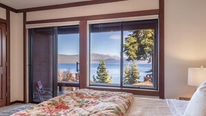 Habitación de hotel con cama, una silla y vistas al mar y las montañas a través de ventanas amplias.