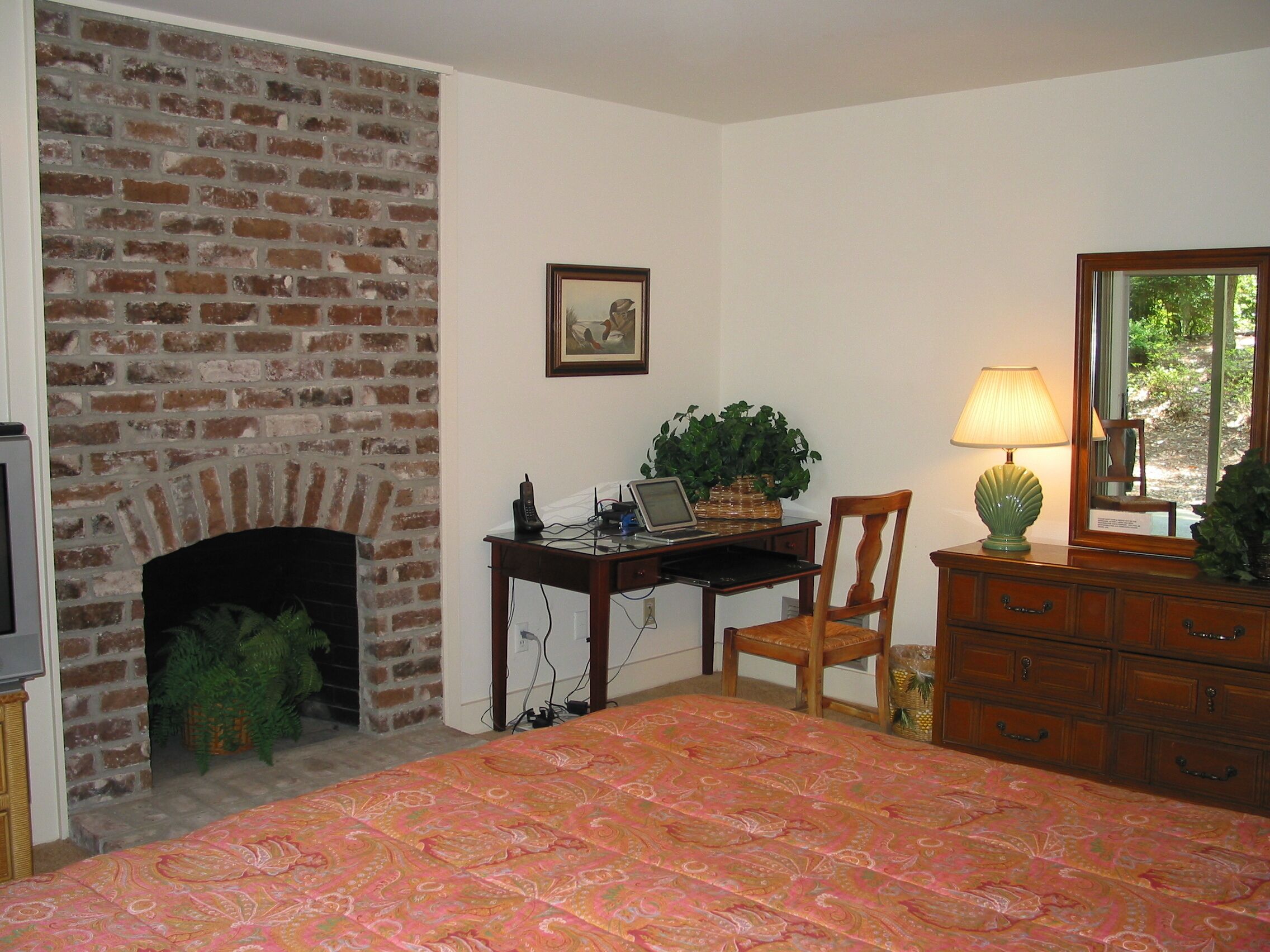 Une chambre avec une cheminée en briques, un bureau avec un ordinateur portable, une chaise, une commode et un lit recouvert d’une couvre-lit à motifs.