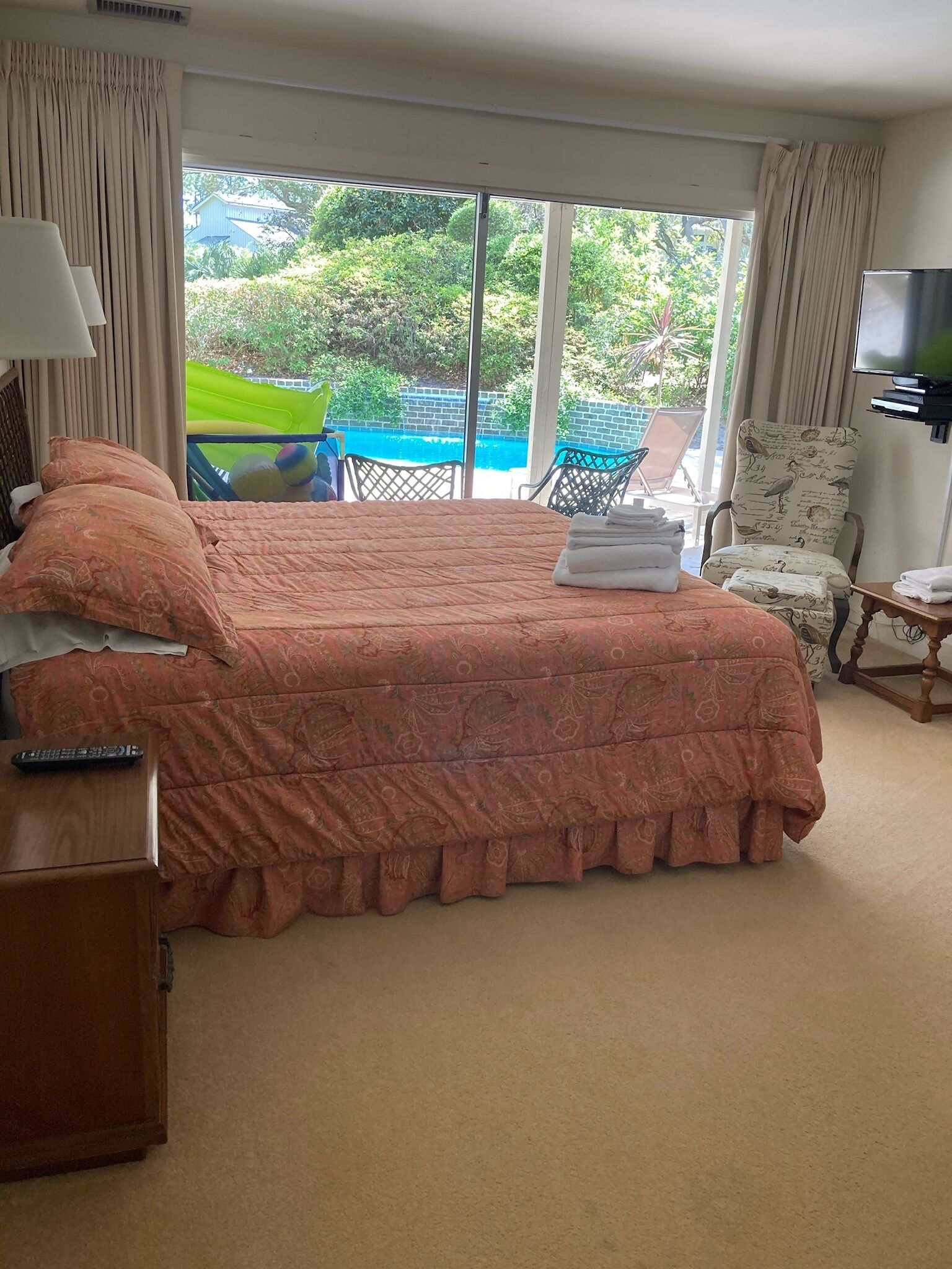 A hotel room with a large bed, a TV, a desk with a remote, and a view of a pool and greenery outside.