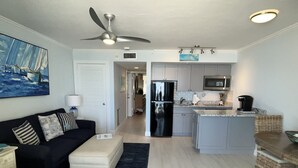 Ein modernes Wohnzimmer mit einem schwarzen Sofa, einem weißen Hocker und einem Gemälde an der Wand. Ein Deckenventilator ist sichtbar, und der Raum geht in eine Küche mit Kühlschrank und Mikrowelle über.