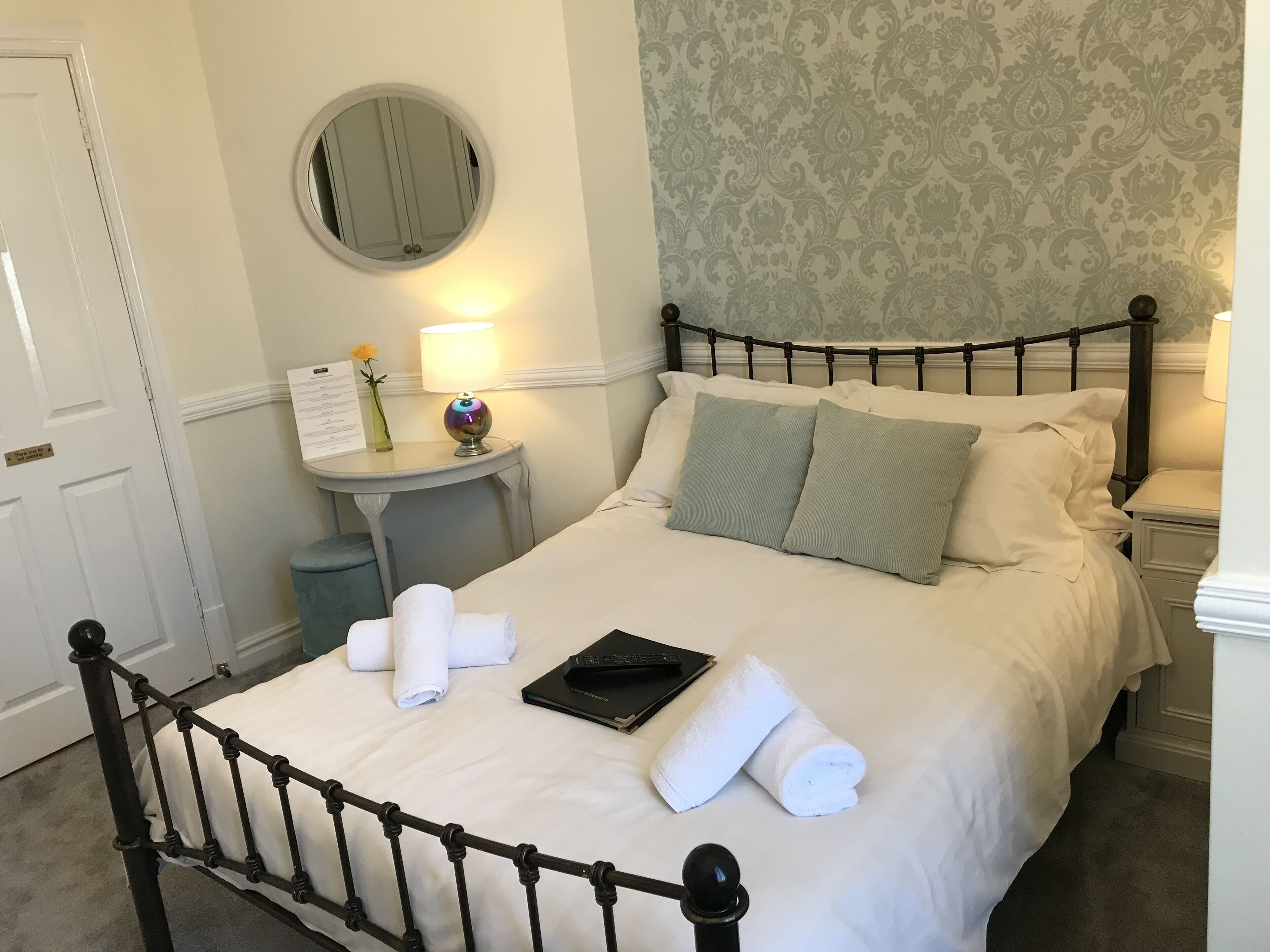 A neatly made bed with white linens, a bedside table with a lamp, a small round mirror, and a floral arrangement.
