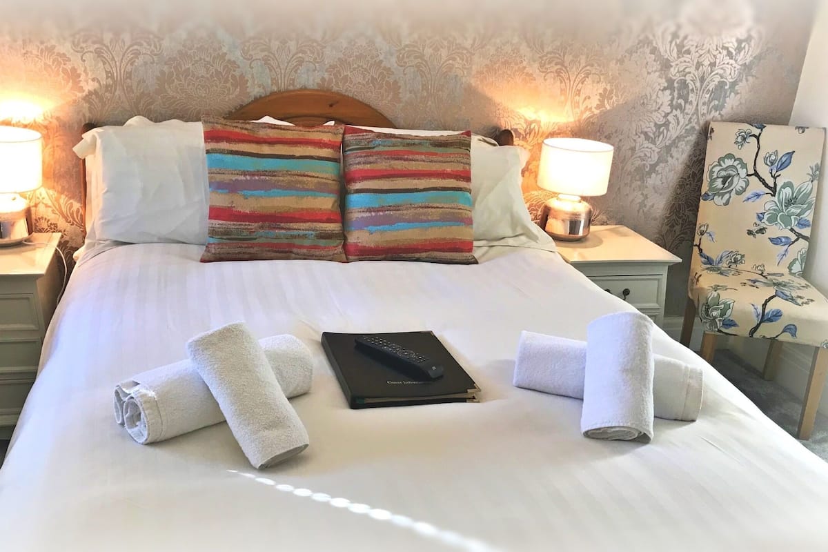 A neatly made bed with white linens, two striped pillows, two bedside lamps, a remote control, and a floral chair.