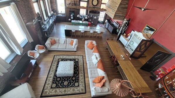 A spacious living area with a large sofa set, a coffee table, and a dining table with chairs. There is a kitchen area with modern appliances and a large clock on the wall. - Indoor Basketball Court, Movie Theatre & Rooftop-Private 3rd floor Penthouse! (Sandusky)
