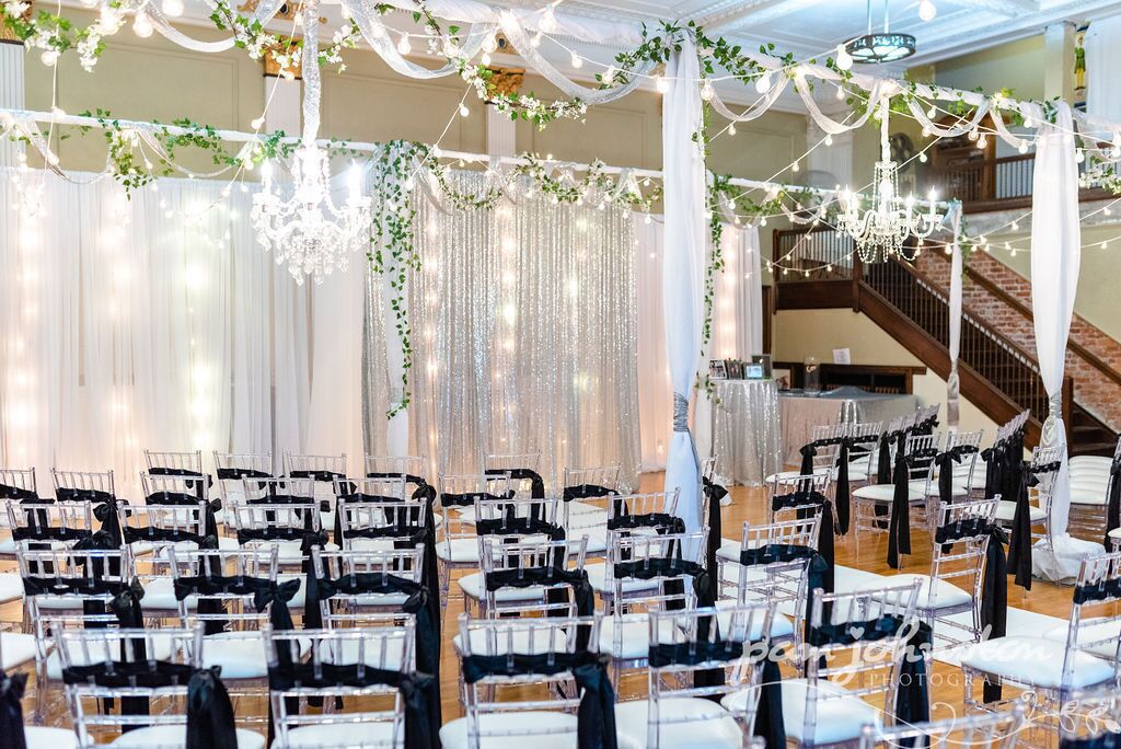 A banquet hall with rows of white chairs, black chair covers, and a chandelier.