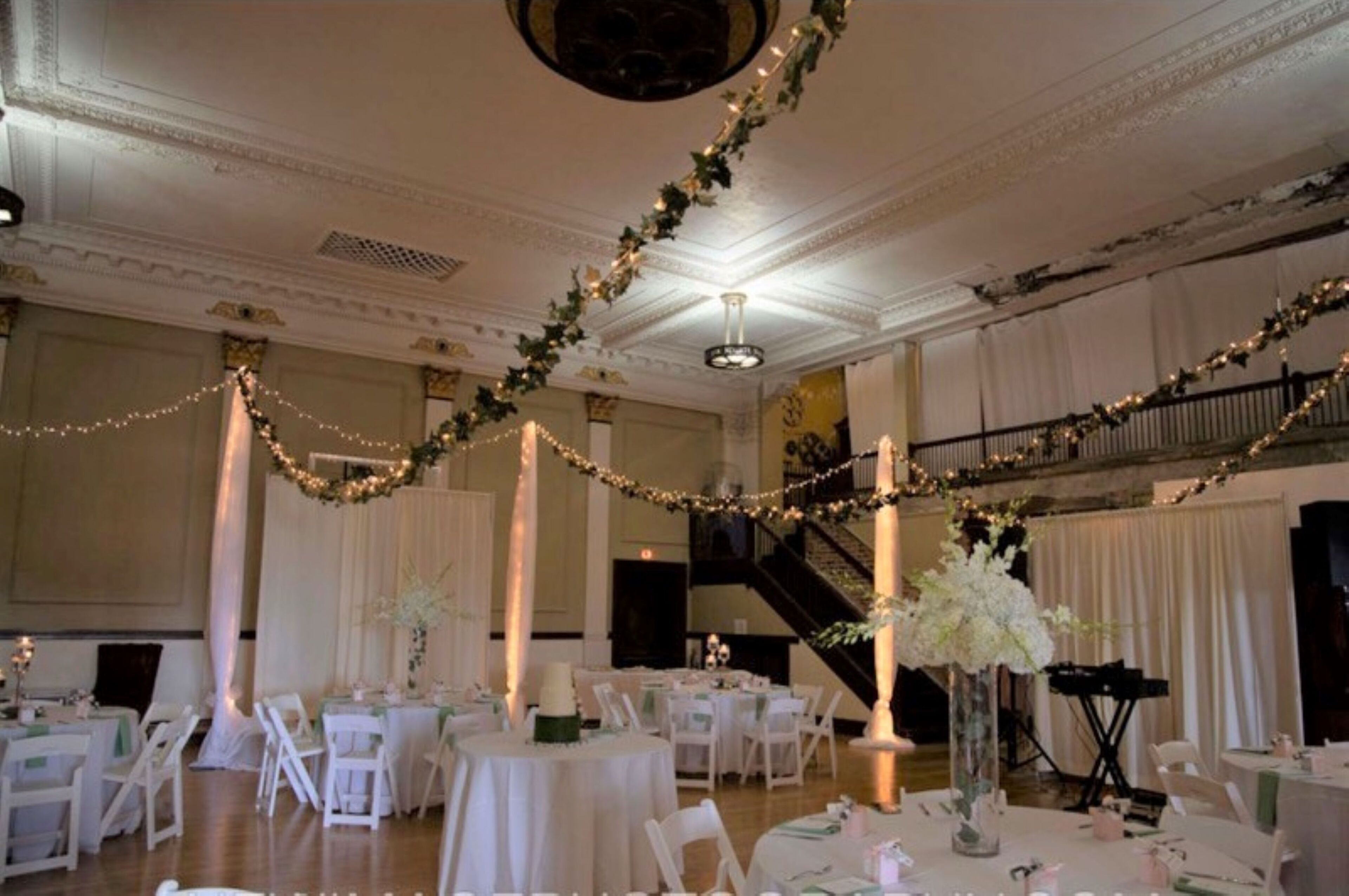 Ein geräumiger Festsaal mit hohen Decken, aufwendigen Stuckarbeiten und einer prächtigen Treppe. Tische sind für eine festliche Veranstaltung gedeckt, und der Raum ist mit Lichterketten und Blumenarrangements dekoriert.