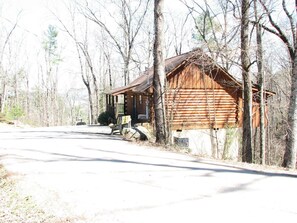 Une cabane rustique en rondins de bois, dotée d’un porche et entourée d’arbres.